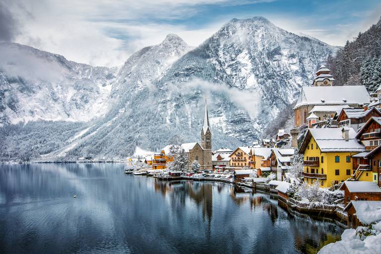 Schneebedecktes Dorf am See mit bunten Häusern und hohem Kirchturm, spiegelnd im ruhigen Wasser unter den imposanten Alpenbergen.