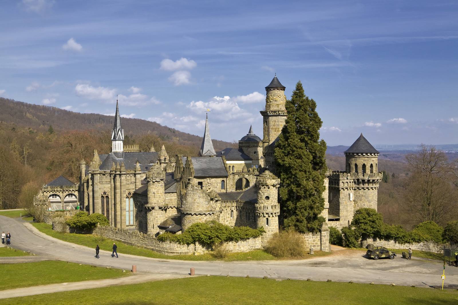 „Ein historisches Steinschloss mit Türmchen und Spitzen vor einer Kulisse aus Bäumen und blauem Himmel, mit einem gewundenen Weg im Vordergrund