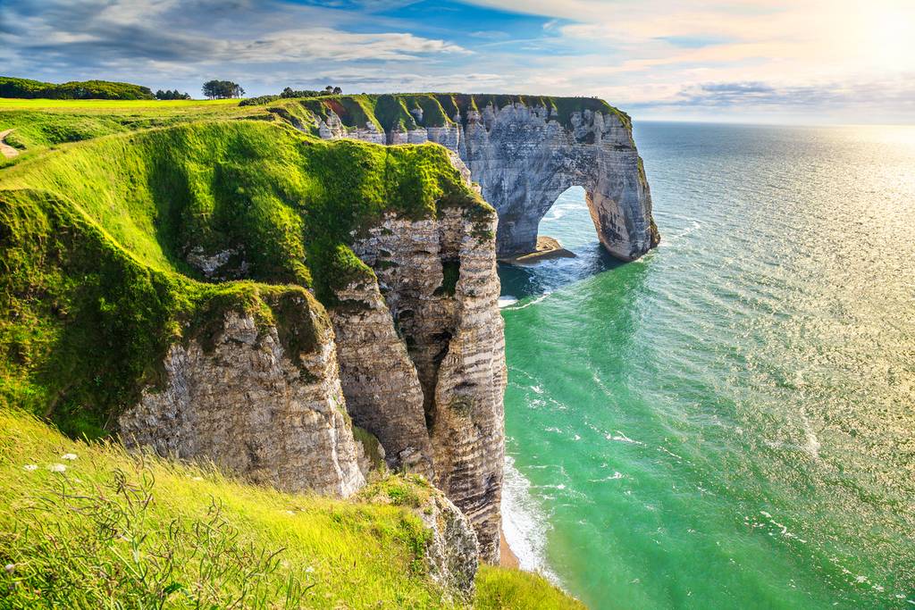 Des falaises au bord de la mer, ornées d'une arche naturelle, recouvertes d'une herbe verdoyante au sommet et baignées par des eaux turquoise, sous un ciel partiellement nuageux.