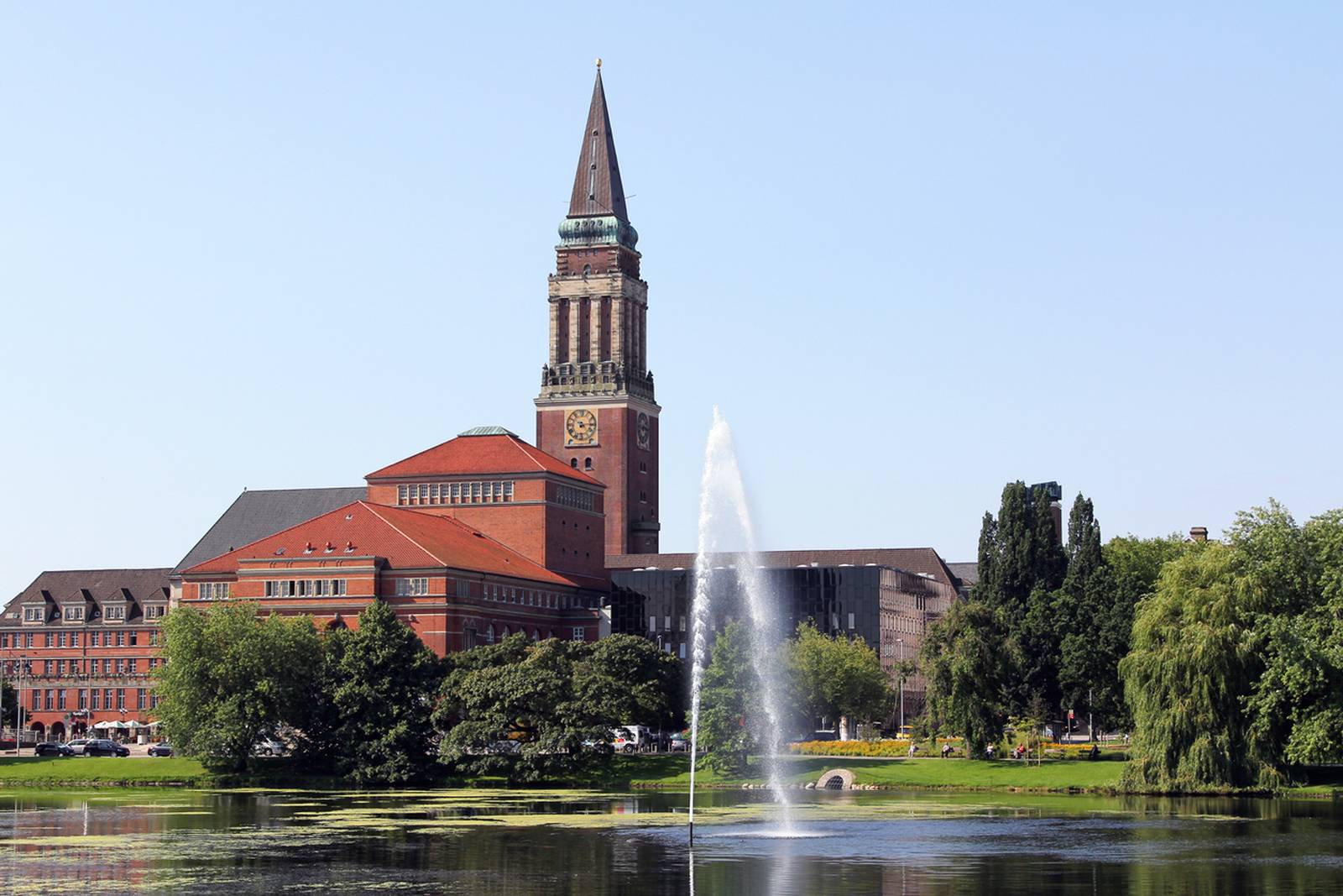 Historisches Gebäude mit hohem Uhrturm an einem See mit Springbrunnen, umgeben von Bäumen unter klarem blauen Himmel
