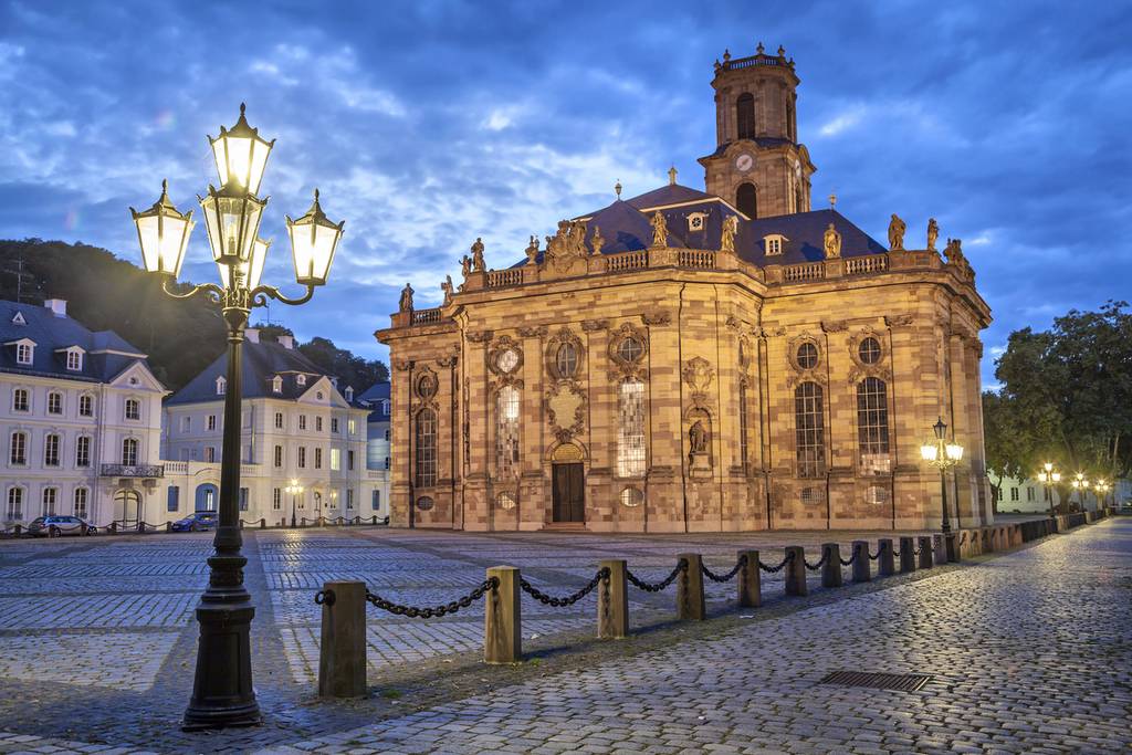 Historische barocke Kirche auf einem Kopfsteinpflasterplatz in der Abenddämmerung, beleuchtet von kunstvoll verzierten Straßenlaternen, vor einem wolkigen Himmel