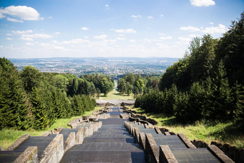 „Blick von der Spitze einer terrassenförmigen Steintreppe, umgeben von üppig grünen Bäumen, mit Aussicht auf eine ferne Stadtsilhouette unter blauem Himmel