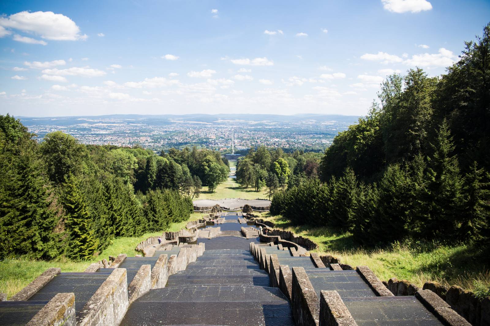 „Blick von der Spitze einer terrassenförmigen Steintreppe, umgeben von üppig grünen Bäumen, mit Aussicht auf eine ferne Stadtsilhouette unter blauem Himmel