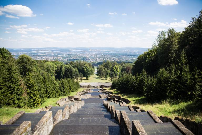 „Blick von der Spitze einer terrassenförmigen Steintreppe, umgeben von üppig grünen Bäumen, mit Aussicht auf eine ferne Stadtsilhouette unter blauem Himmel
