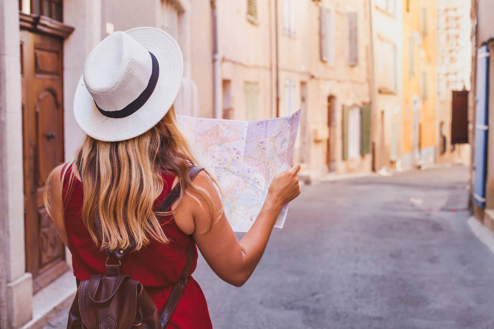 Een vrouw met een hoed en een rode jurk houdt een landkaart in haar hand en staat in een smal Europees straatje dat wordt omzoomd door pastelkleurige gebouwen.