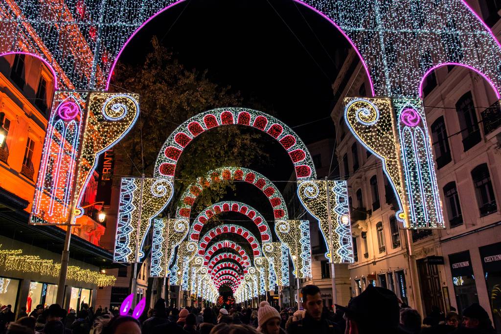 Beleuchtete Torbögen mit bunten Lichtern säumen eine belebte Straße und sorgen für eine festliche Atmosphäre, während Menschen nachts darunter entlangschlendern.