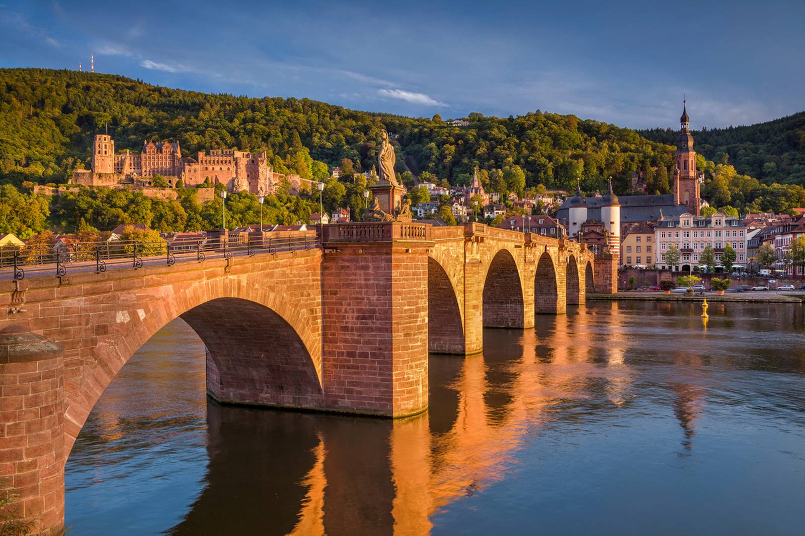 Alte Steinbrücke über einem ruhigen Fluss mit Spiegelungen, umgeben von üppigen Hügeln und historischen Gebäuden unter klarem blauen Himmel beim Sonnenuntergang