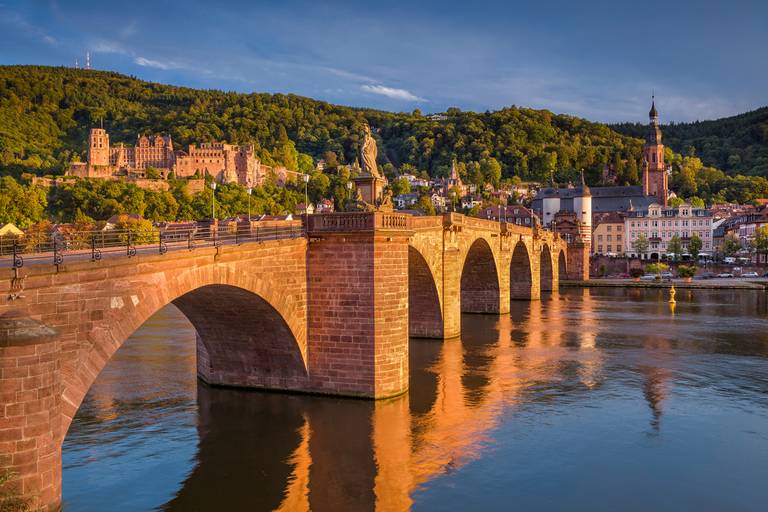Alte Steinbrücke über einem ruhigen Fluss mit Spiegelungen, umgeben von üppigen Hügeln und historischen Gebäuden unter klarem blauen Himmel beim Sonnenuntergang