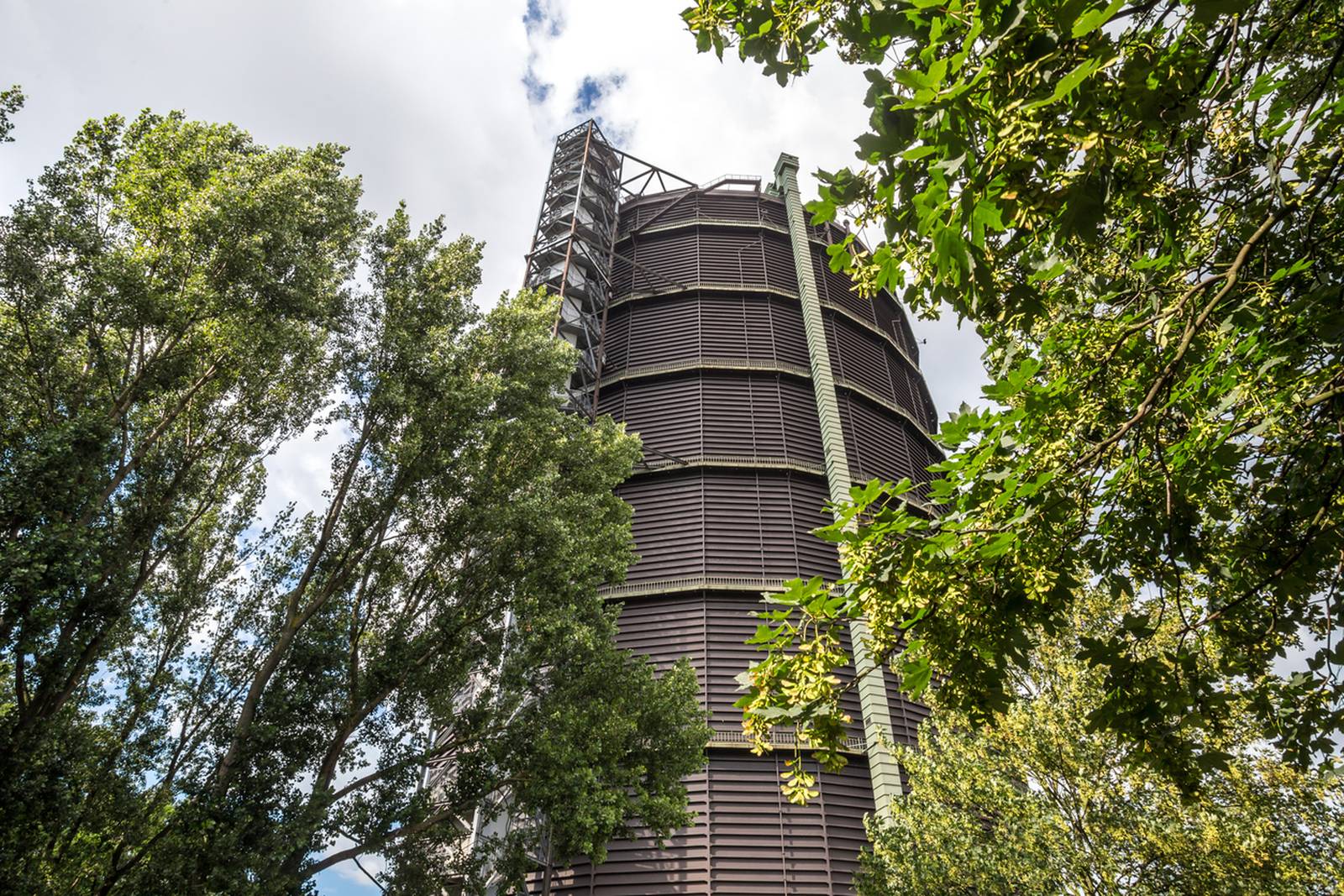 Hohes, zylindrisches Industriegebäude, umgeben von grünen Bäumen, unter einem teilweise bewölkten Himmel