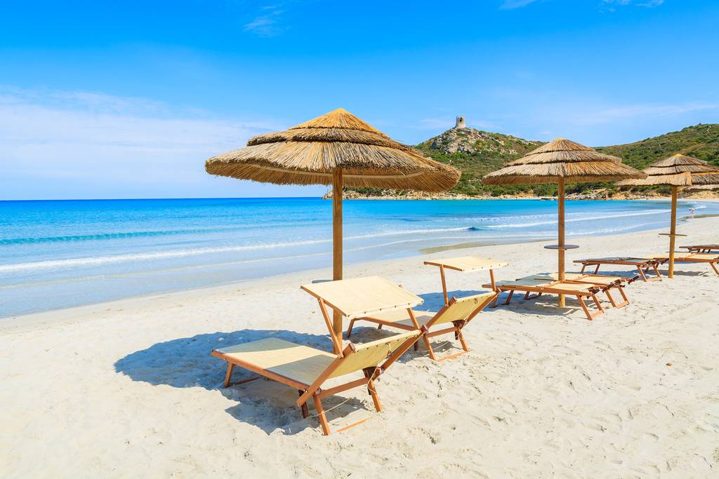 Sonniger Strand mit Strohmsonnenschirmen und Liegen auf weißem Sand. Ruhiges, klares blaues Meer und ein entfernter Turm auf einem grünen Hügel unter strahlend blauem Himmel.