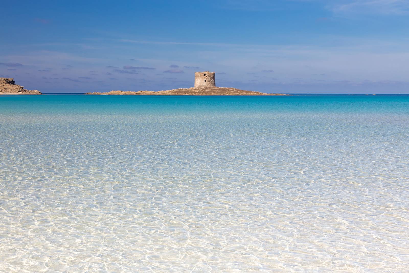 Klares türkisfarbenes Wasser und eine felsige Insel mit historischem Steinturm unter strahlend blauem Himmel.