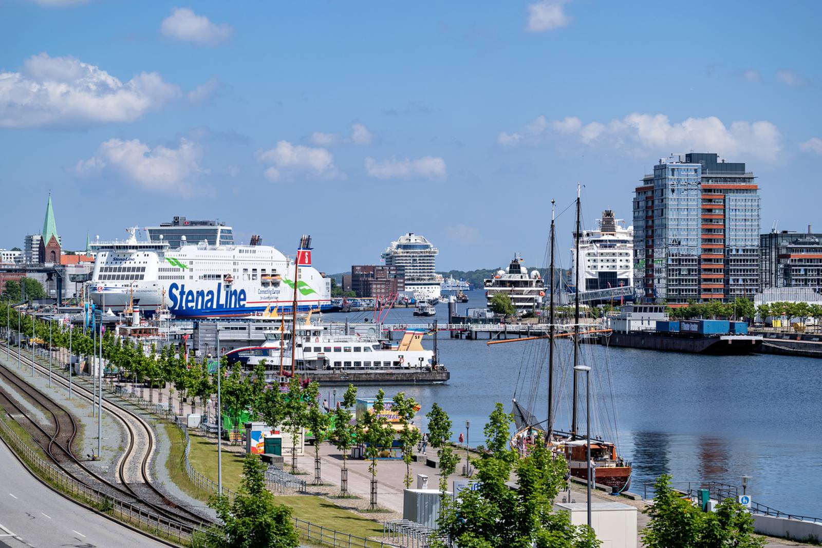 Lebhafte Hafensilhouette mit Fähren, modernen Gebäuden und einer baumbestandenen Uferpromenade unter klarem blauen Himmel