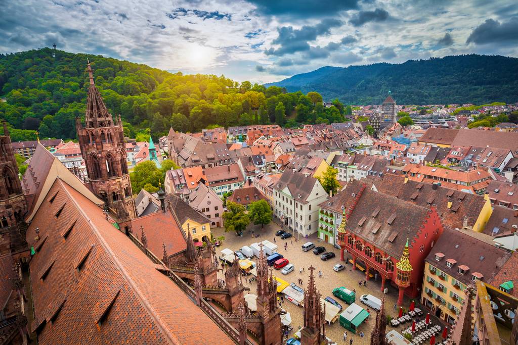 Luftaufnahme einer historischen europäischen Stadt mit bunten Häusern, einem Dom und grünen Hügeln unter einem teilweise bewölkten Himmel