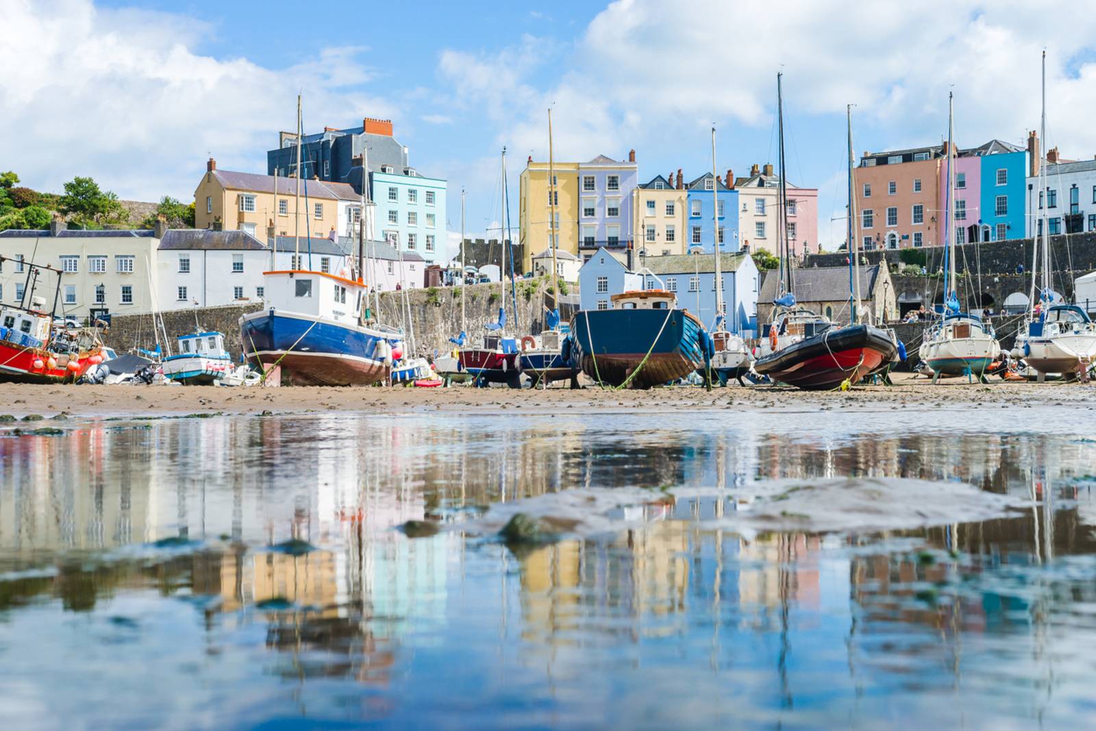 Colorful boats and pastel buildings reflect in calm waters at the  harbor of tenby, under a partly cloudy sky.