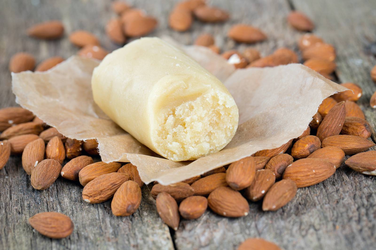 Eine Rolle Marzipan auf Backpapier, umgeben von ganzen Mandeln auf einer rustikalen Holzoberfläche