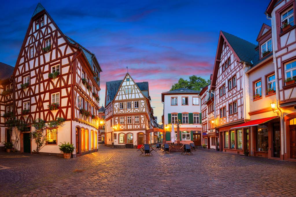 Bezaubernder europäischer Dorfplatz in der Dämmerung mit Fachwerkhäusern, Kopfsteinpflaster und warmem Straßenlicht unter einem farbenfrohen Abendhimmel