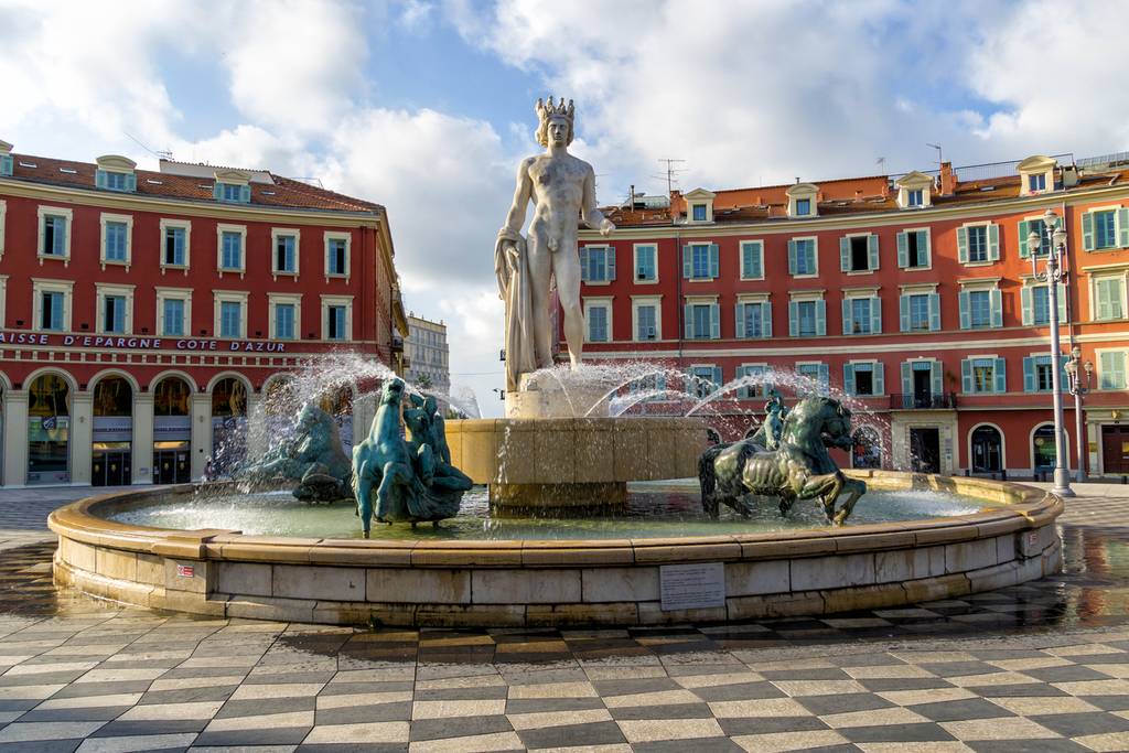 Brunnen mit einer zentralen Statue und umliegenden Pferdeskulpturen auf einem Platz, umgeben von roten Gebäuden unter einem teilweise bewölkten Himmel