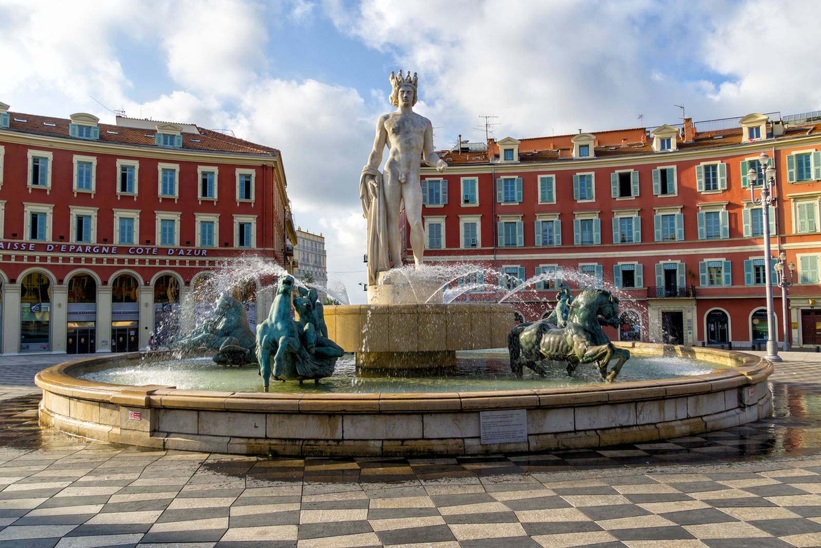 Brunnen mit einer zentralen Statue und umliegenden Pferdeskulpturen auf einem Platz, umgeben von roten Gebäuden unter einem teilweise bewölkten Himmel