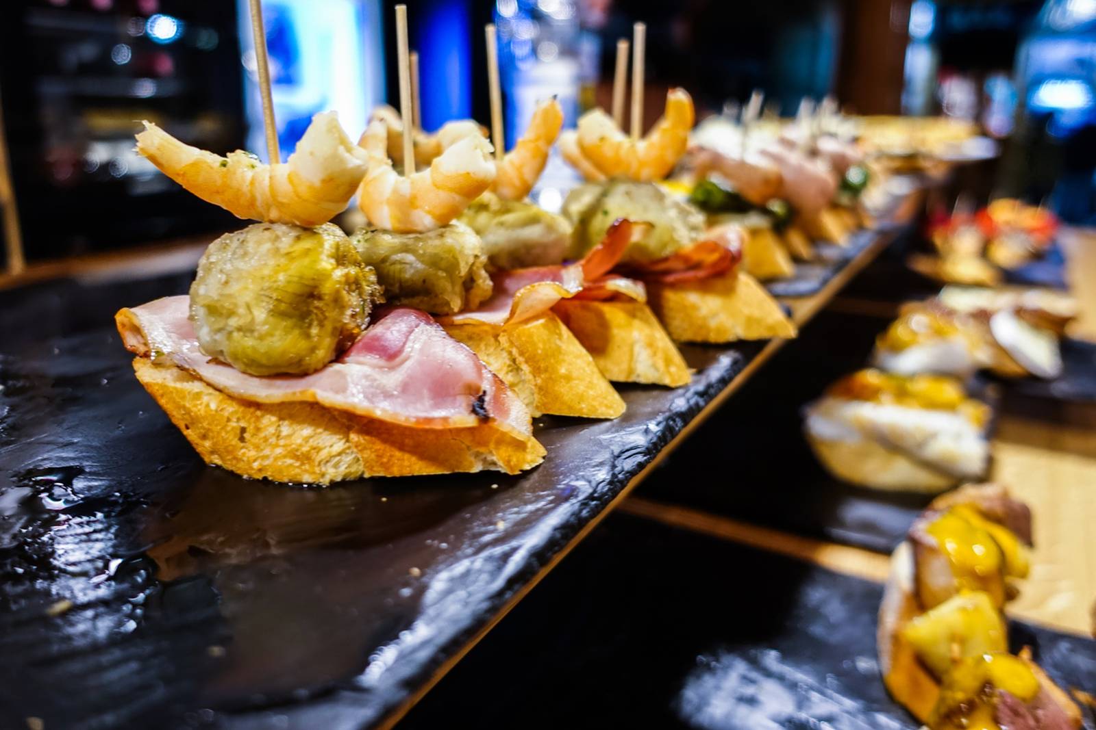 Bilbao: Close-up of assorted tapas on a black slate, featuring shrimp, ham, and vegetables on sliced bread, displayed in a row.