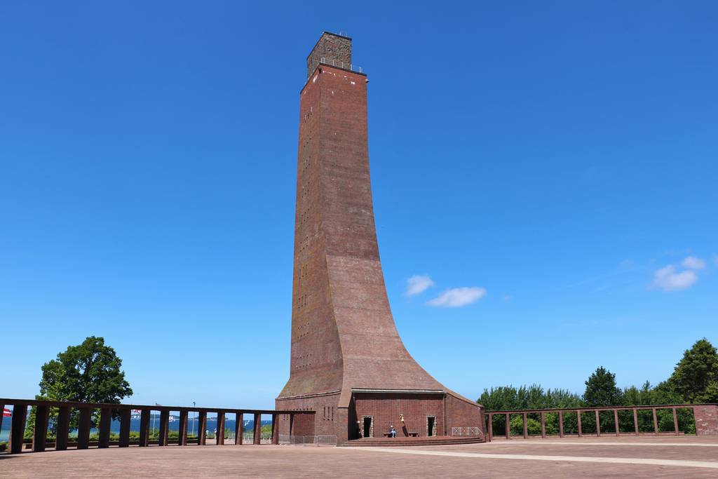 Hoher, backsteinerner Marine-Gedenkturm mit geschwungenem Sockel unter klarem blauen Himmel, umgeben von Bäumen und einer niedrigen Mauer