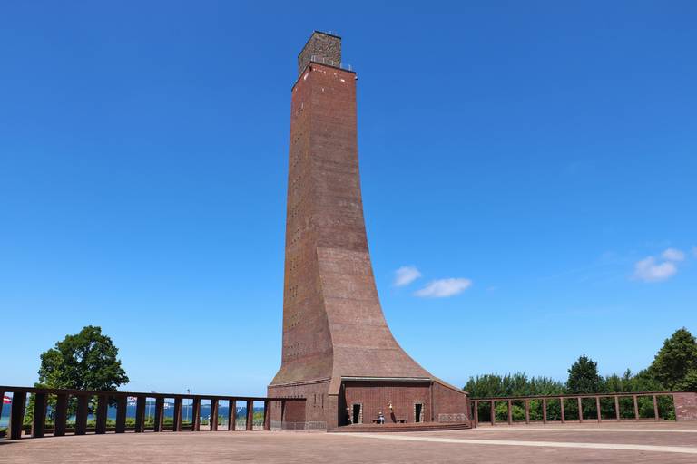Hoher, backsteinerner Marine-Gedenkturm mit geschwungenem Sockel unter klarem blauen Himmel, umgeben von Bäumen und einer niedrigen Mauer