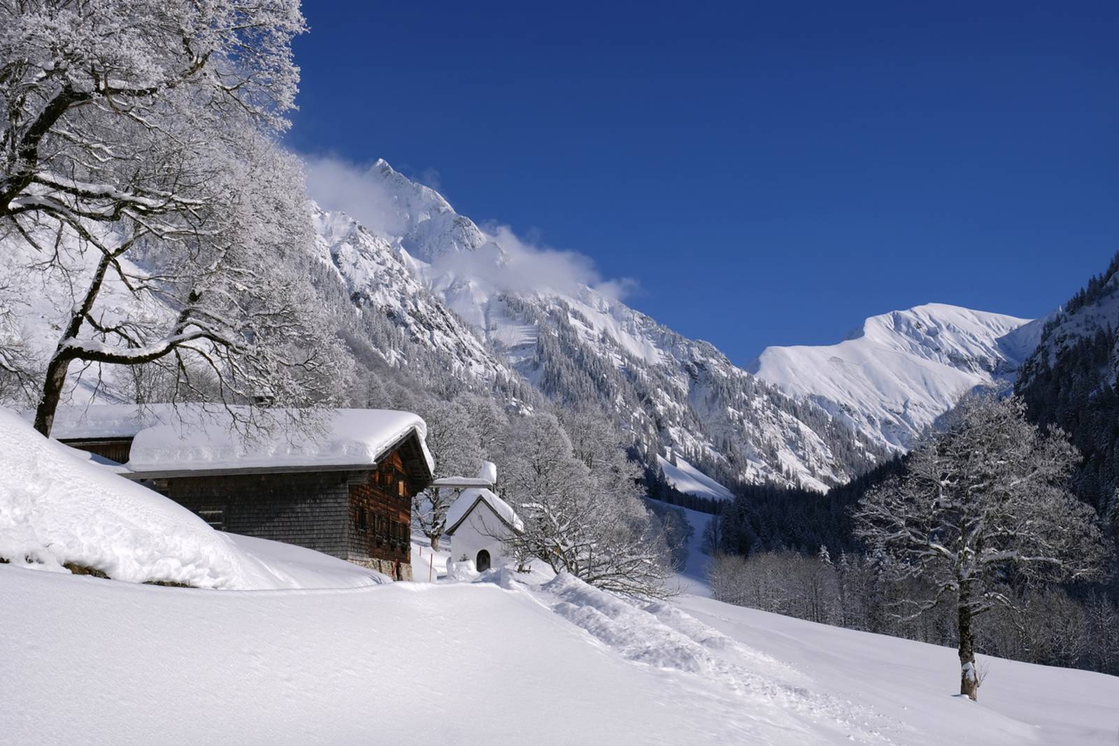 Schneebedeckte Chalets und Bäume in einem sonnenbeschienenen Bergtal, umgeben von imposanten verschneiten Gipfeln unter einem klaren, tiefblauen Himmel.