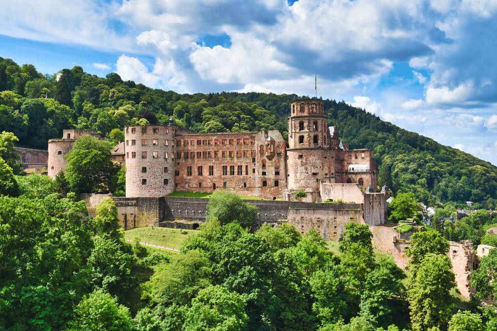 Heidelberger Schloss auf einem üppig bewachsenen Hügel unter einem wolkigen blauen Himmel, umgeben von grünen Wäldern.