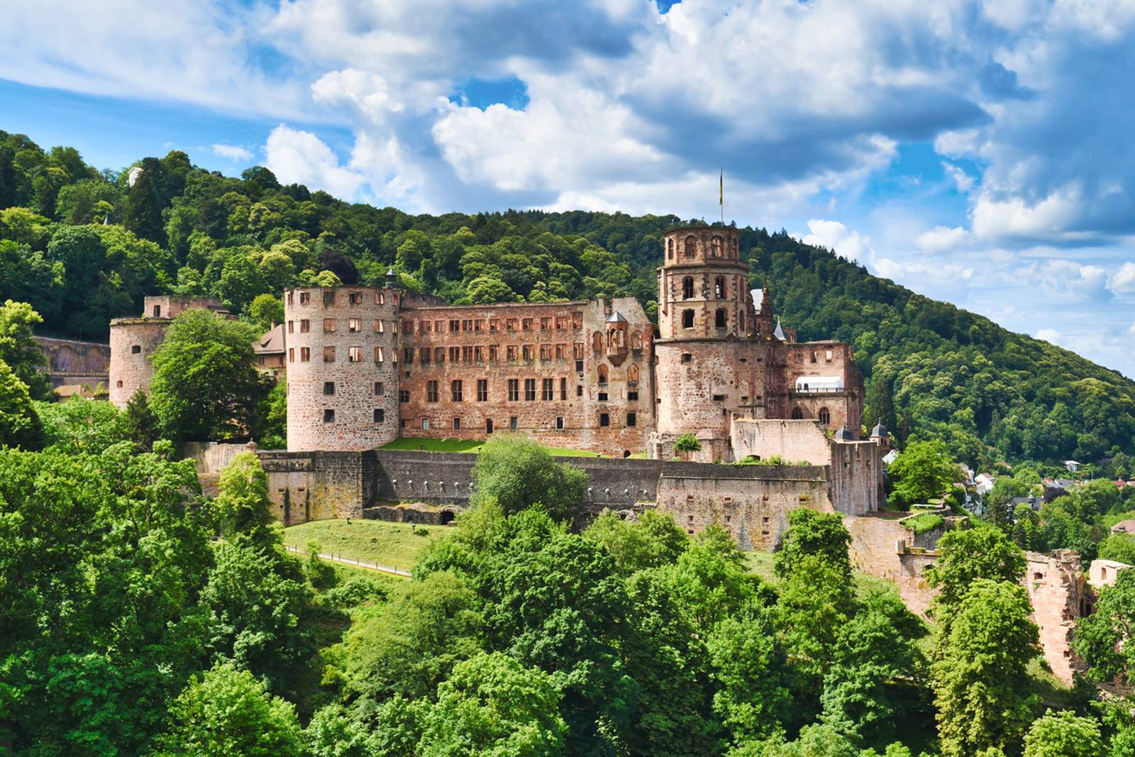 Das Heidelberger Schloss, umgeben von üppigem Grün, vor der Kulisse eines teils bewölkten Himmels und bewaldeter Hügel
