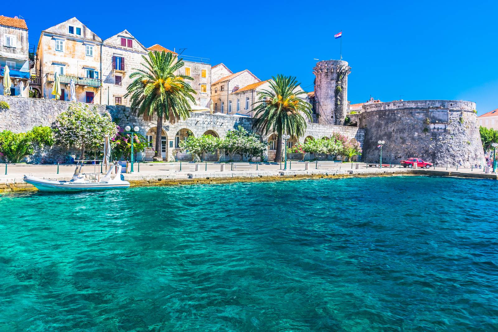 Scenic view of a Korcula with stone buildings, palm trees, and a fortress by clear turquoise water under a bright blue sky.