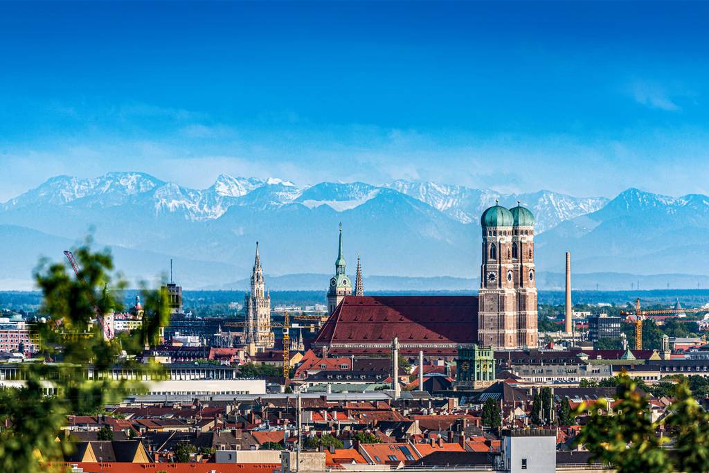 Skyline of Munich with red-roofed buildings, prominent church towers, and snow-capped mountains in the background under a clear blue sky.
