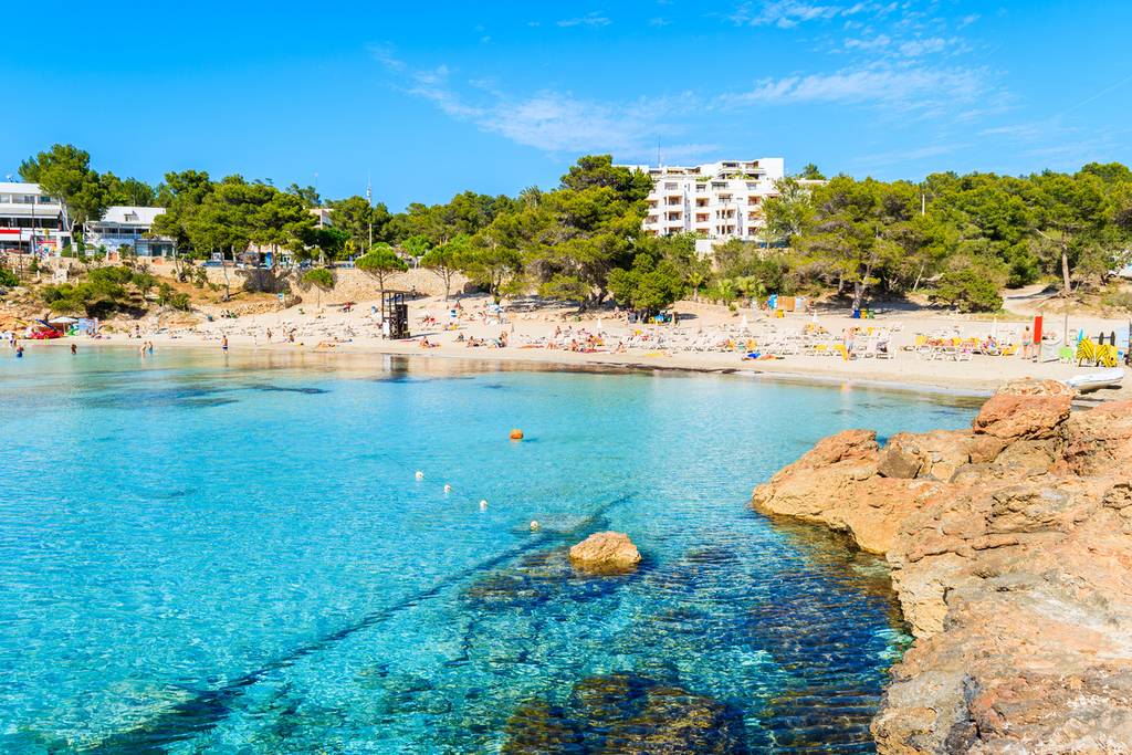 Baia dalle acque turchesi e cristalline a Portinat con spiaggia di sabbia, bagnanti, costa rocciosa e spiagge circondate da alberi sotto un cielo blu intenso.