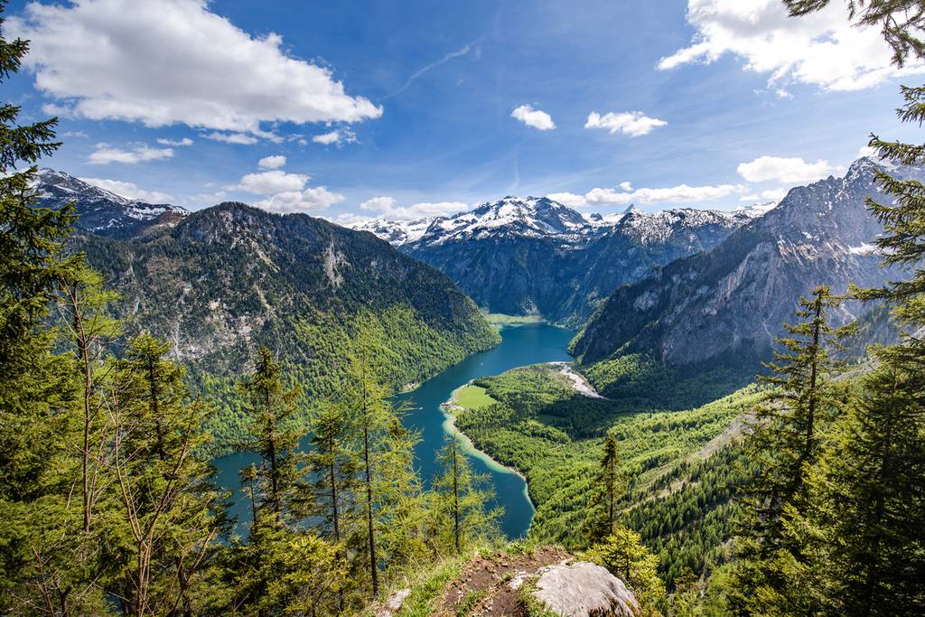 Panoramic view of a turquoise alpine lake winding between forested valleys and snow-capped mountains under a blue sky.