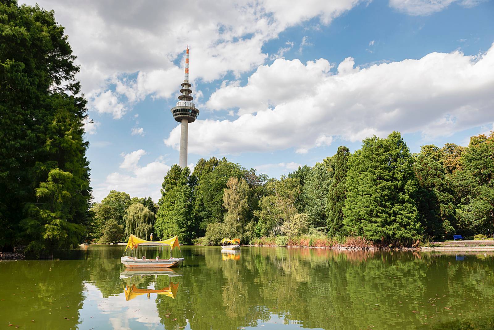 Ruhiger See mit einem kleinen Boot, umgeben von üppigen Bäumen. Im Hintergrund ragt ein hoher Turm unter einem leicht bewölkten Himmel empor.