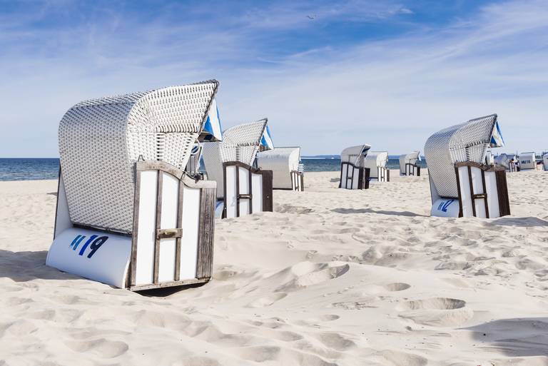 Geflochtene Strandkörbe an einem sandigen Ufer unter blauem Himmel, mit dem Meer im Hintergrund, die eine ruhige Küstenszene schaffen.