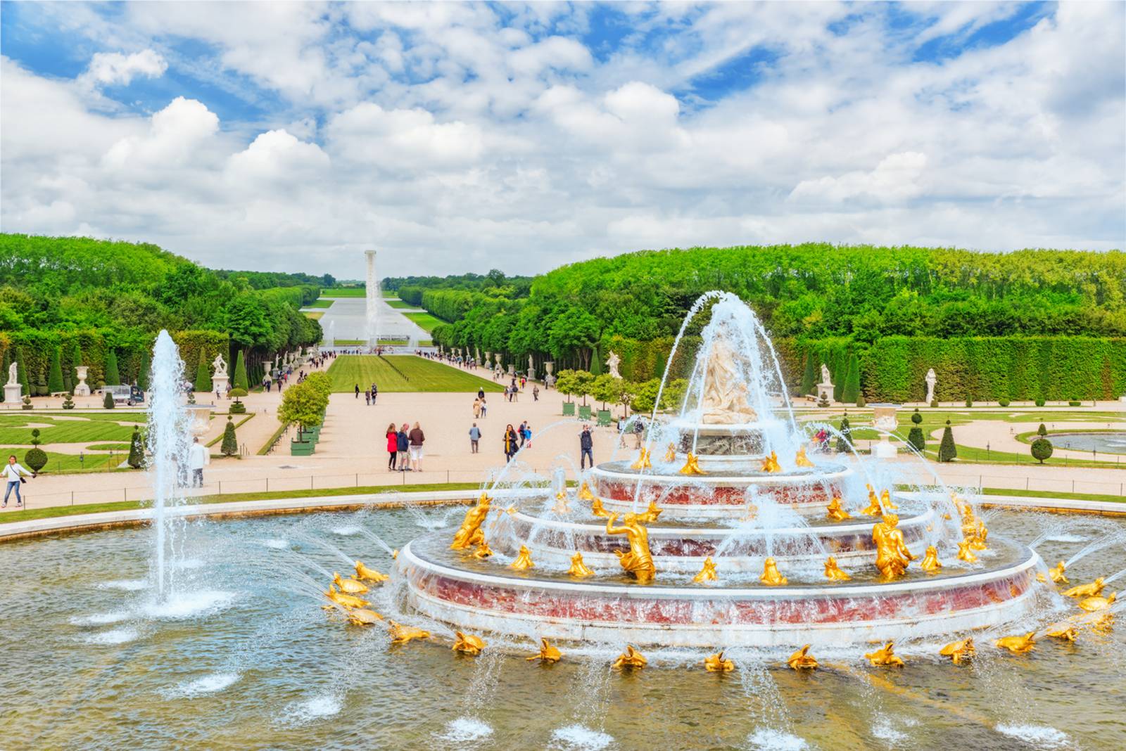 Fountain with golden statues in a landscaped garden, surrounded by greenery and a wide pathway with people walking under a partly cloudy sky.