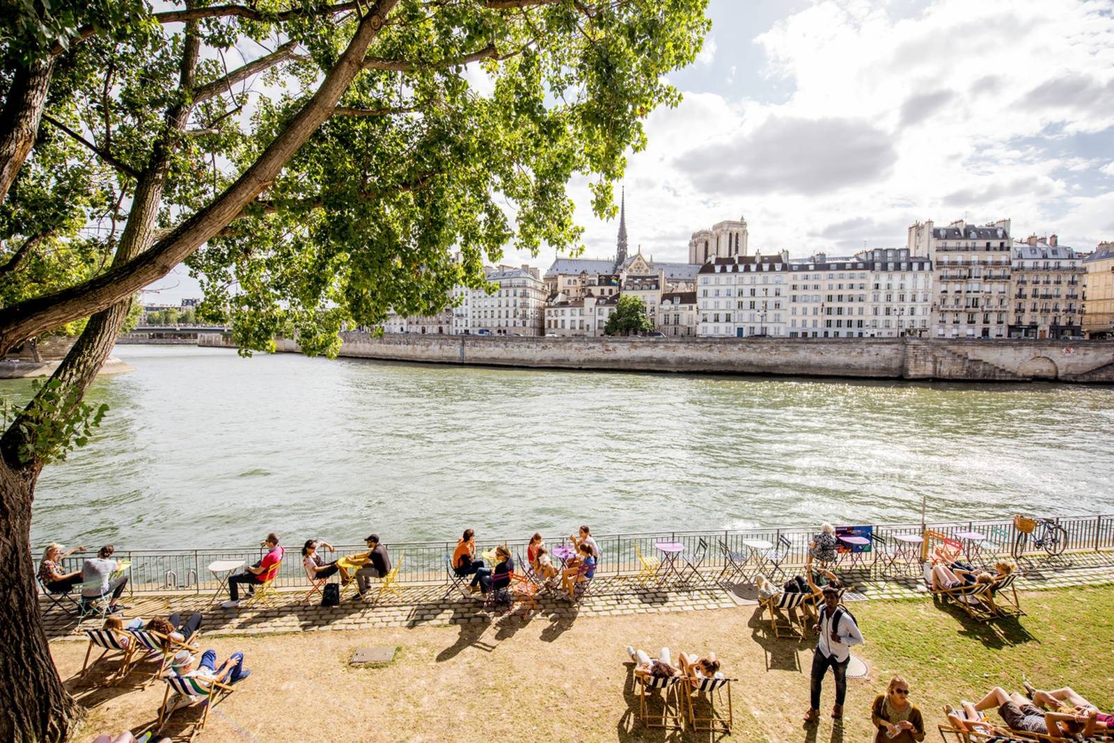 People relax on chairs and benches along the Seine River under trees, with historic buildings in the background on a sunny day.