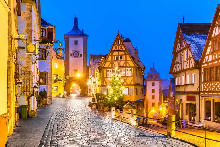 Kopfsteinpflasterstraße in Rothenburg ob der Tauber, Deutschland, mit Fachwerkhäusern und einem beleuchteten Weihnachtsbaum in der Dämmerung.