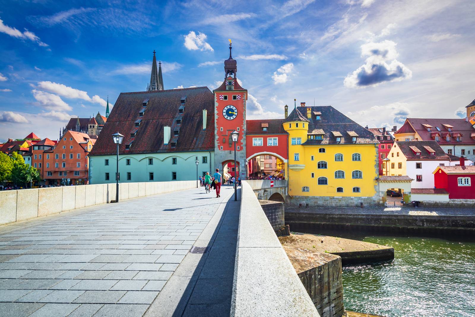 Bunte historische Gebäude säumen eine Straße am Flussufer in Regensburg, Deutschland, mit einem markanten Uhrturm unter einem teilweise bewölkten Himmel