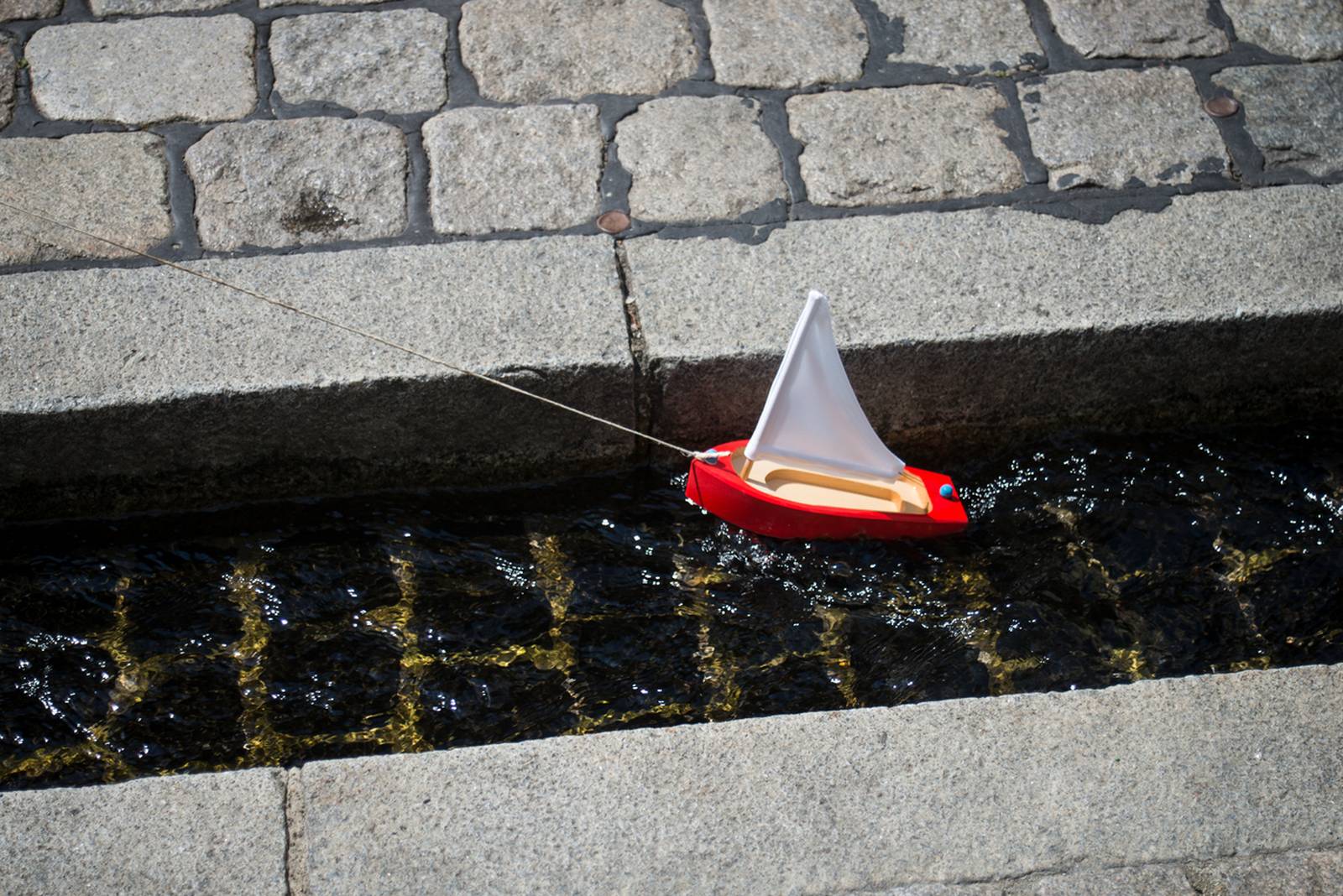 Ein kleines rotes Spielzeug-Segelboot mit weißem Segel treibt an einem sonnigen Tag in einem schmalen, mit Steinen eingefassten Wasserkanal