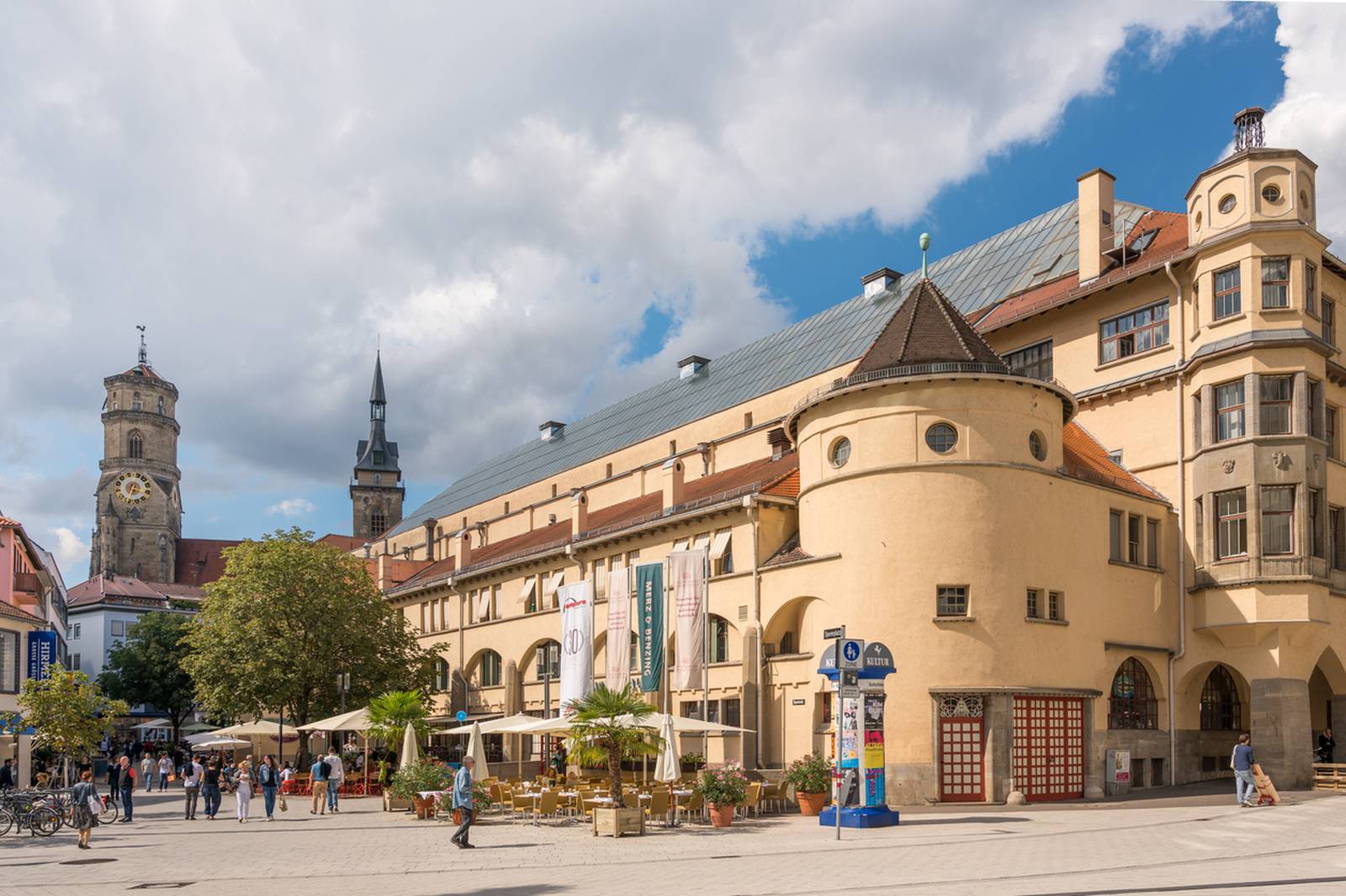 Historisches Gebäude mit rundem Turm auf einem sonnigen Platz, umgeben von Menschen und Straßencafés; Kirchtürme sind im Hintergrund sichtbar