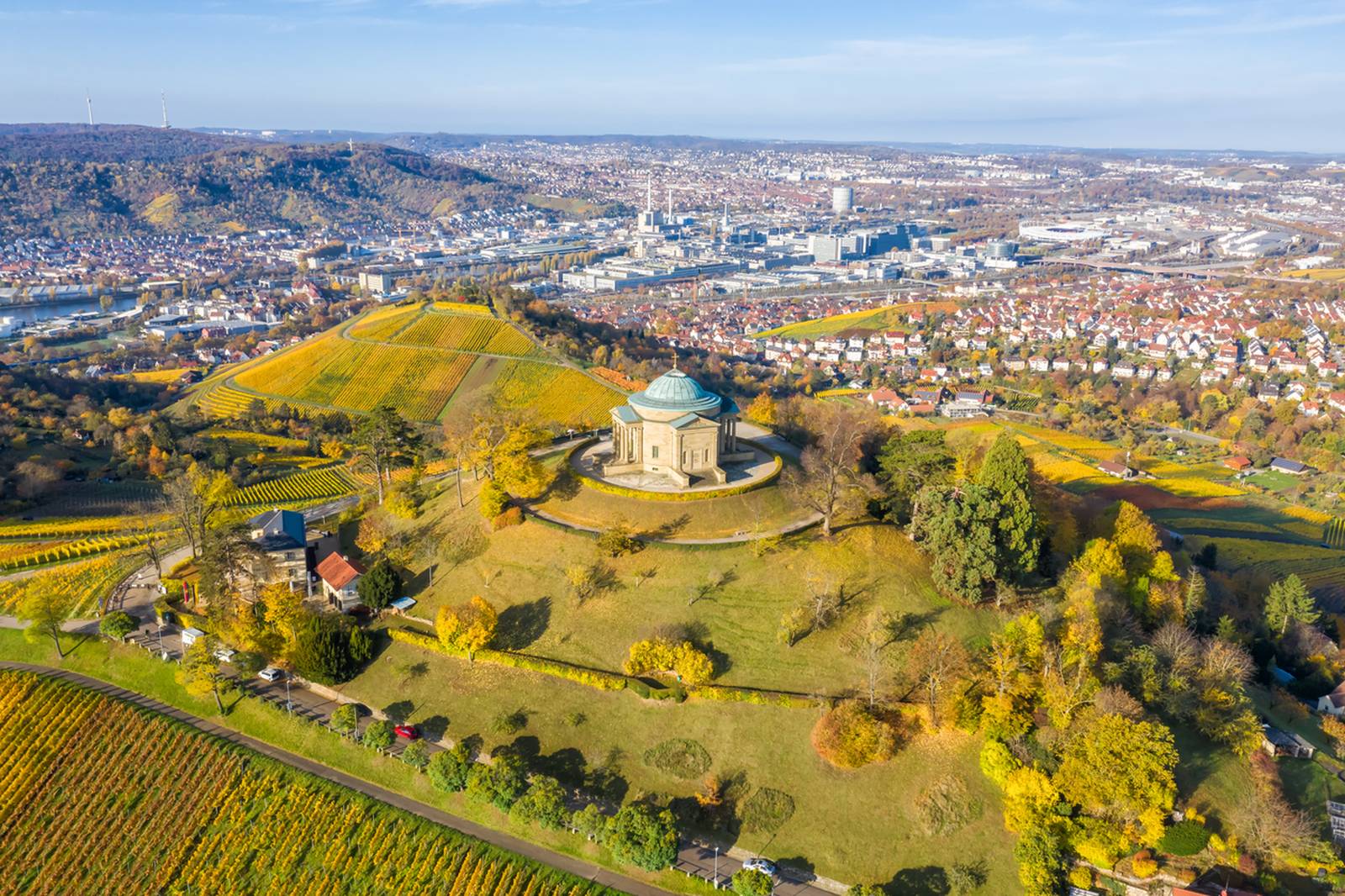 Luftaufnahme eines Gebäudes auf einem Hügel, umgeben von Weinbergen und Bäumen, mit Blick auf eine Stadtlandschaft unter klarem blauem Himmel