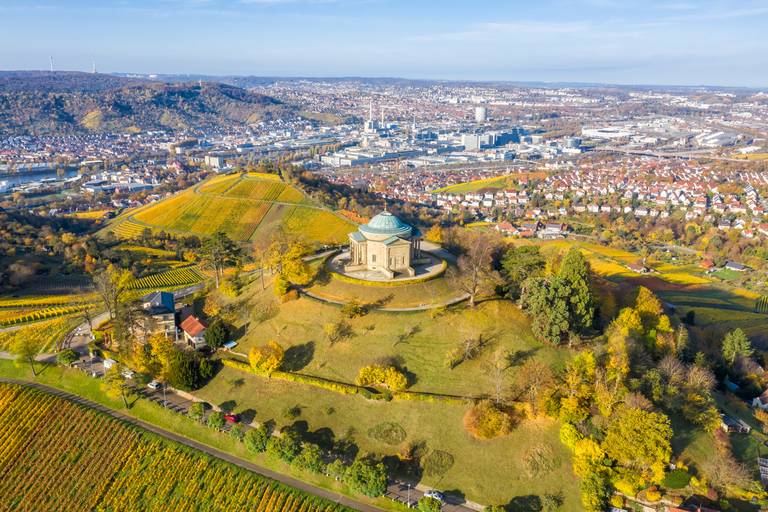 Luftaufnahme eines Gebäudes auf einem Hügel, umgeben von Weinbergen und Bäumen, mit Blick auf eine Stadtlandschaft unter klarem blauem Himmel
