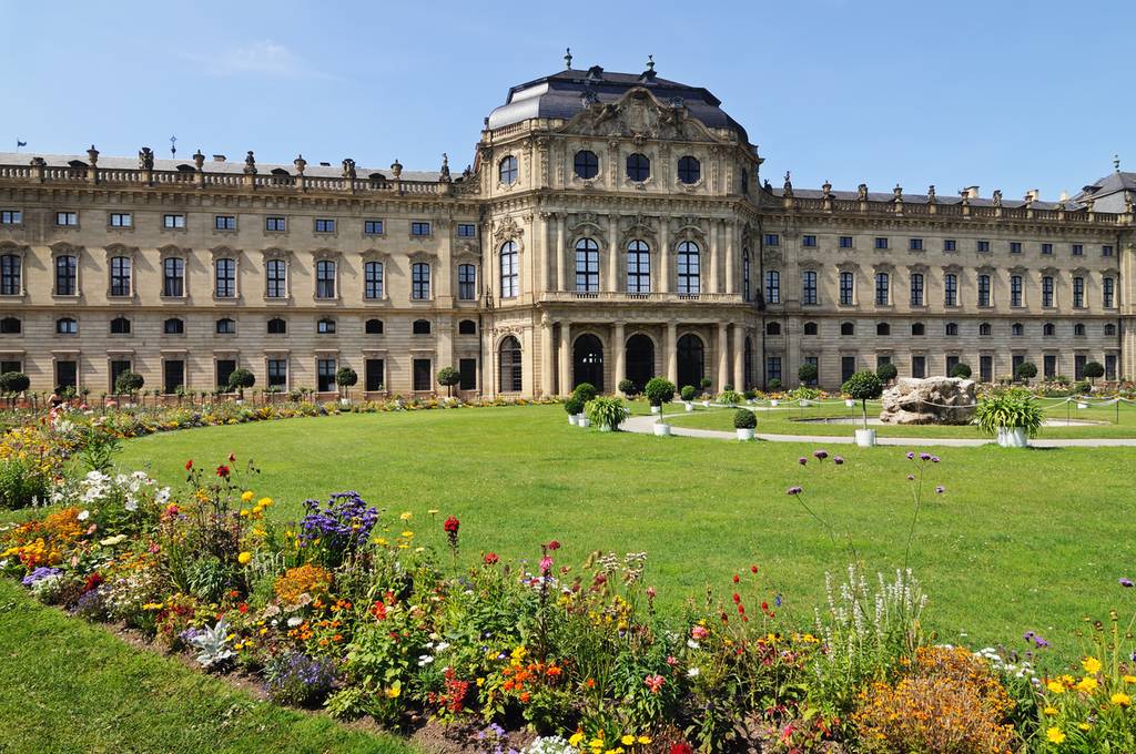 Historisches barockes Schloss mit kunstvoll verzierter Fassade, weitläufigem grünen Rasen und farbenfrohen Blumenbeeten unter klarem blauen Himmel