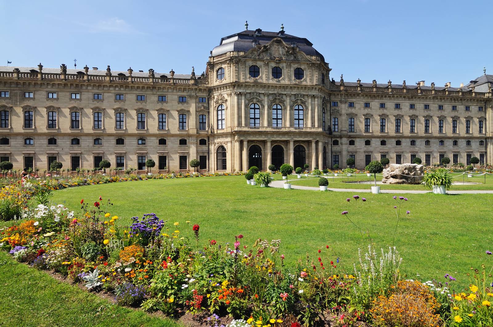 Historisches barockes Schloss mit kunstvoll verzierter Fassade, weitläufigem grünen Rasen und farbenfrohen Blumenbeeten unter klarem blauen Himmel