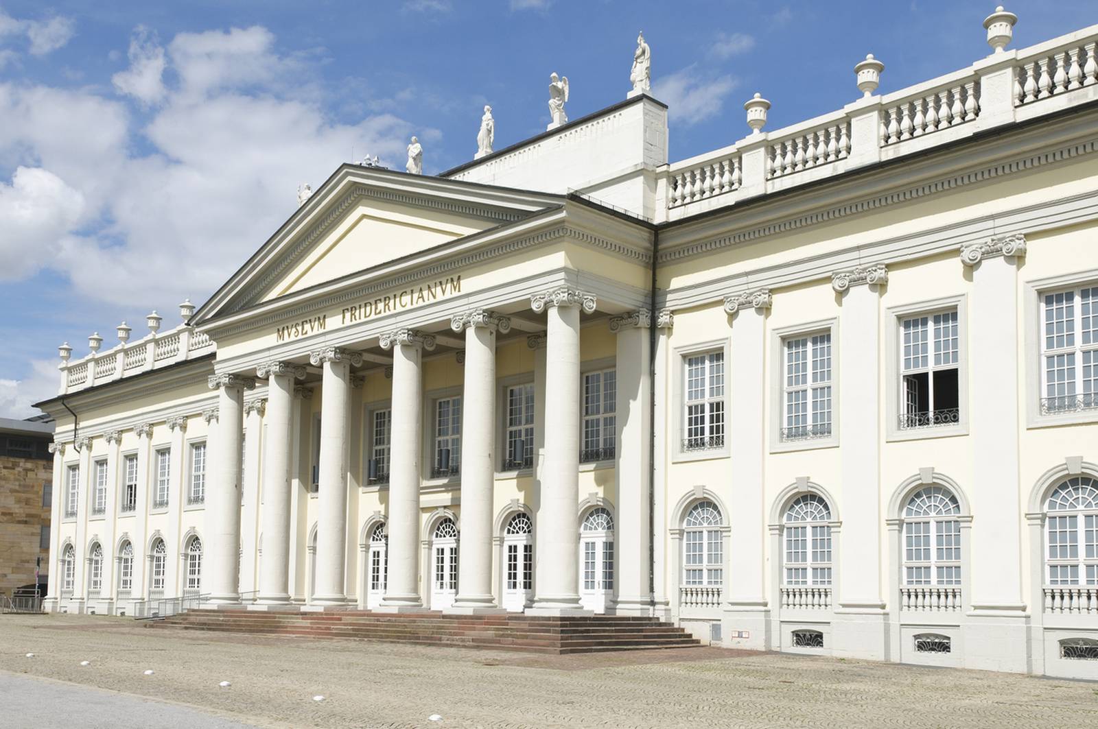 Neoklassizistisches Gebäude mit hohen Säulen, verzierten Fenstern und Statuen auf dem Dach, vor einem blauen Himmel mit vereinzelten Wolken