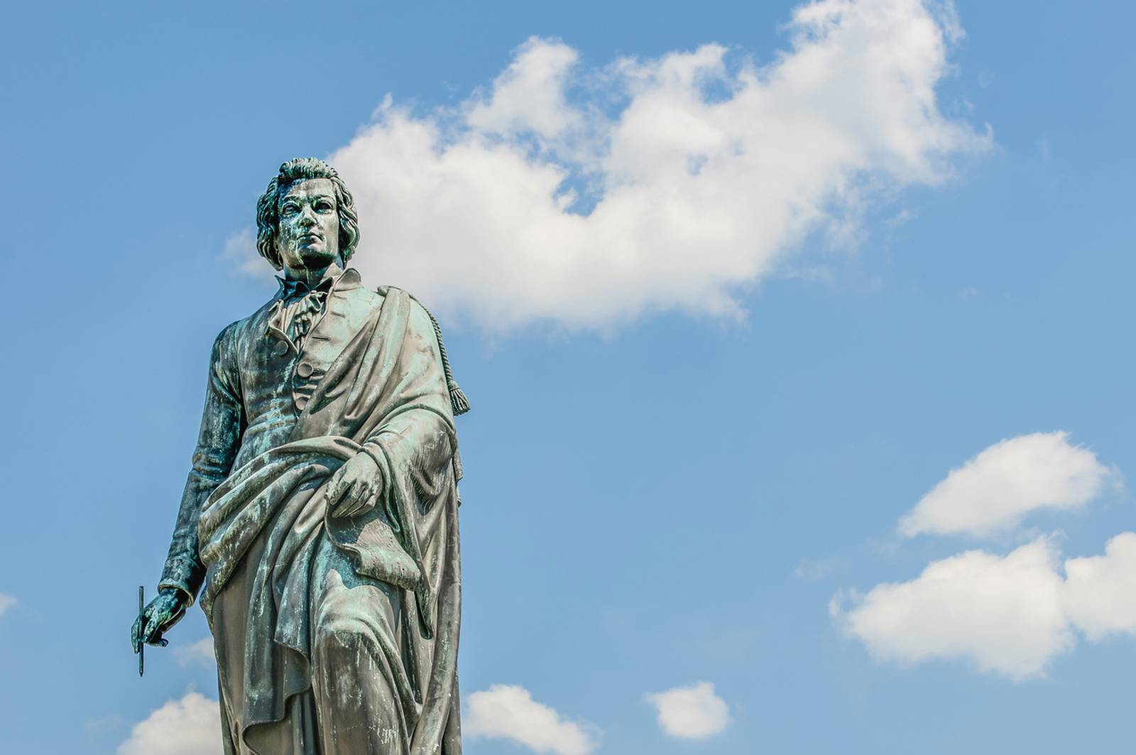 Statue einer historischen Persönlichkeit mit einer Schriftrolle in der Hand, vor blauem Himmel mit vereinzelten Wolken