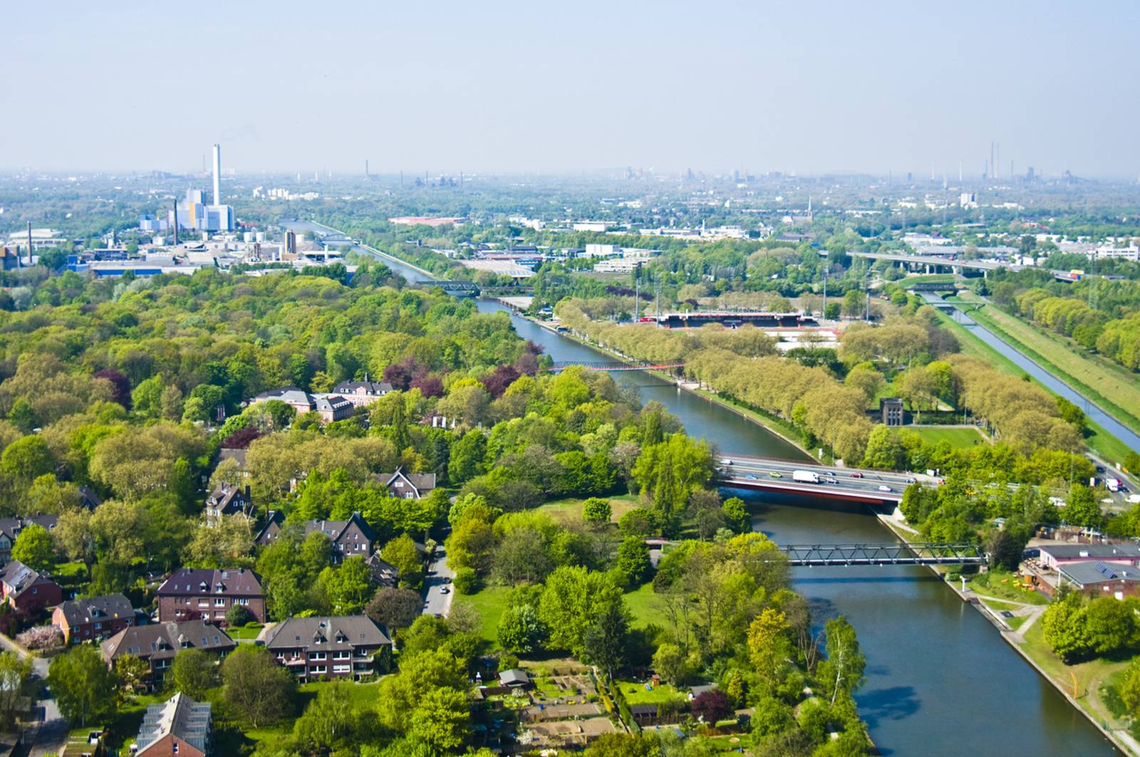 Luftaufnahme einer üppig grünen Landschaft mit einem Fluss, Brücken, Wohngebieten und Industriegebäuden unter klarem blauen Himmel