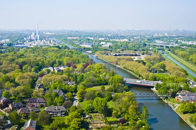 Luftaufnahme einer üppig grünen Landschaft mit einem Fluss, Brücken, Wohngebieten und Industriegebäuden unter klarem blauen Himmel