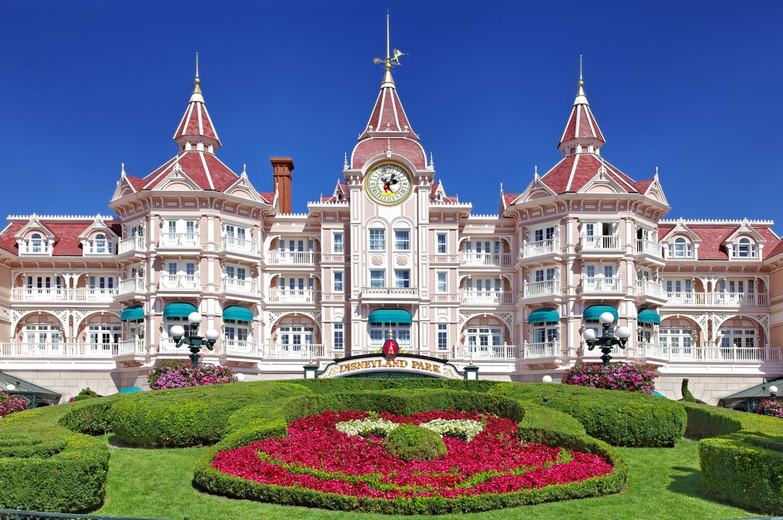 Disneyland paris, Victorian-style building with red roofs, clock tower, and manicured gardens featuring a Mickey Mouse floral design, under a clear blue sky.