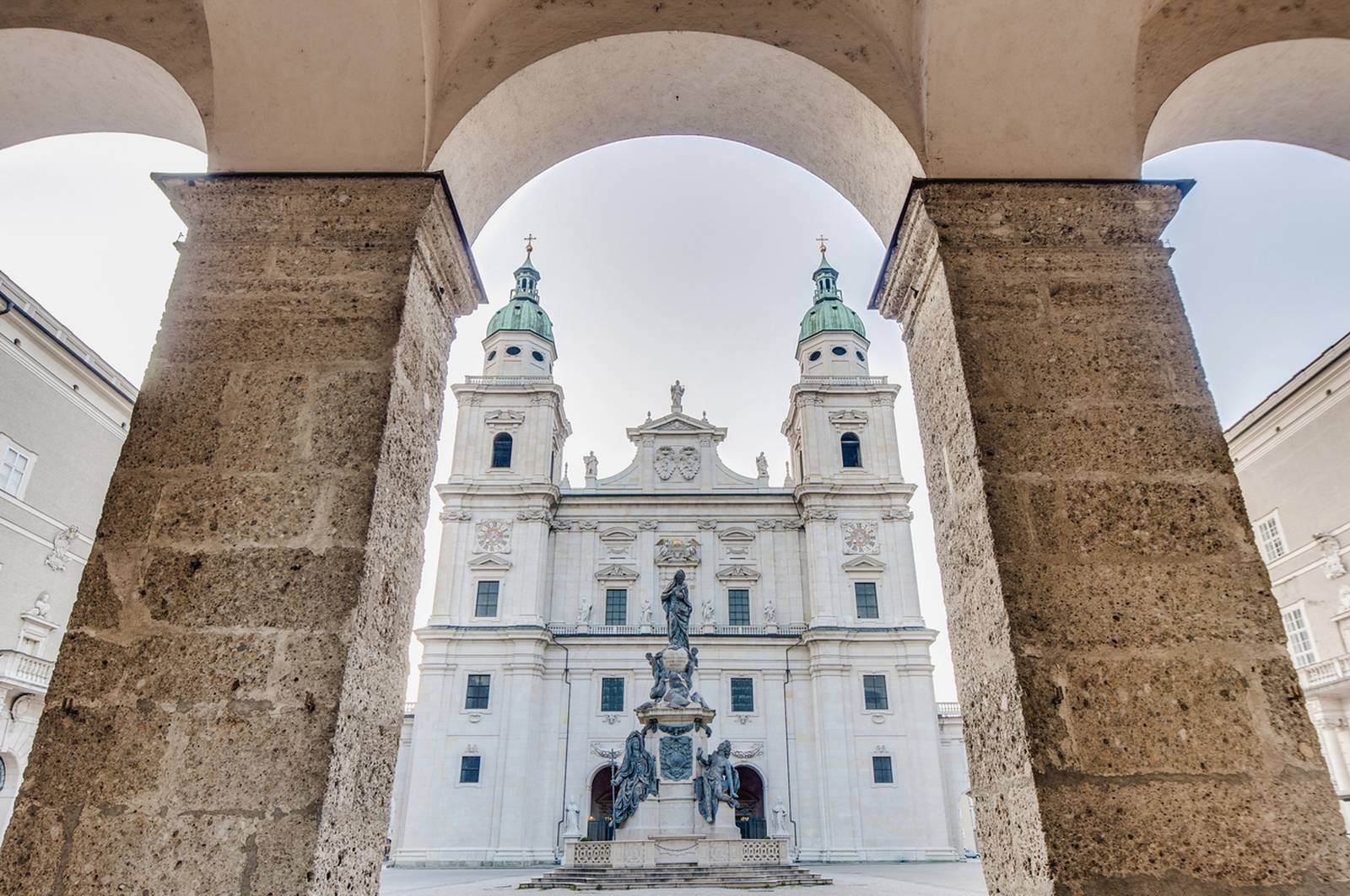 Blick auf eine historische Kathedrale mit zwei grün­kuppeligen Türmen, eingerahmt von steinernen Bögen, mit einer zentralen Statue im Vordergrund